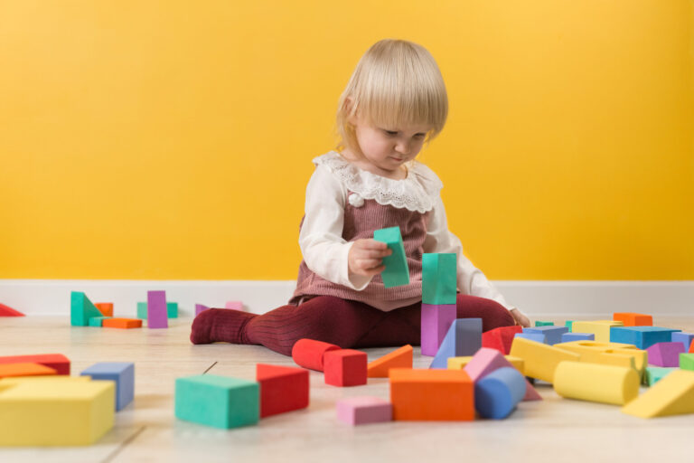 A little focused girl is sitting on the floor in a room and is building a tower of multi-colored blocks of a constructor