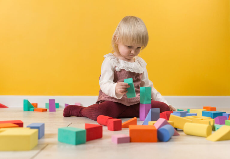 A little focused girl is sitting on the floor in a room and is building a tower of multi-colored blocks of a constructor